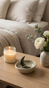Rustic ceramic ring dish with crescent moon on wooden nightstand with lit candle, flowers and linen bedding