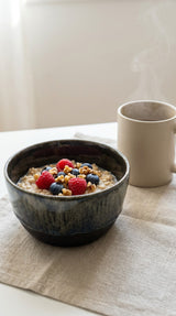 Moody dark ceramic bowl filled with oatmeal, berries and walnuts next to steaming mug