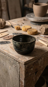 Handmade dark ceramic serving bowl on pottery workbench surrounded by tools and clay